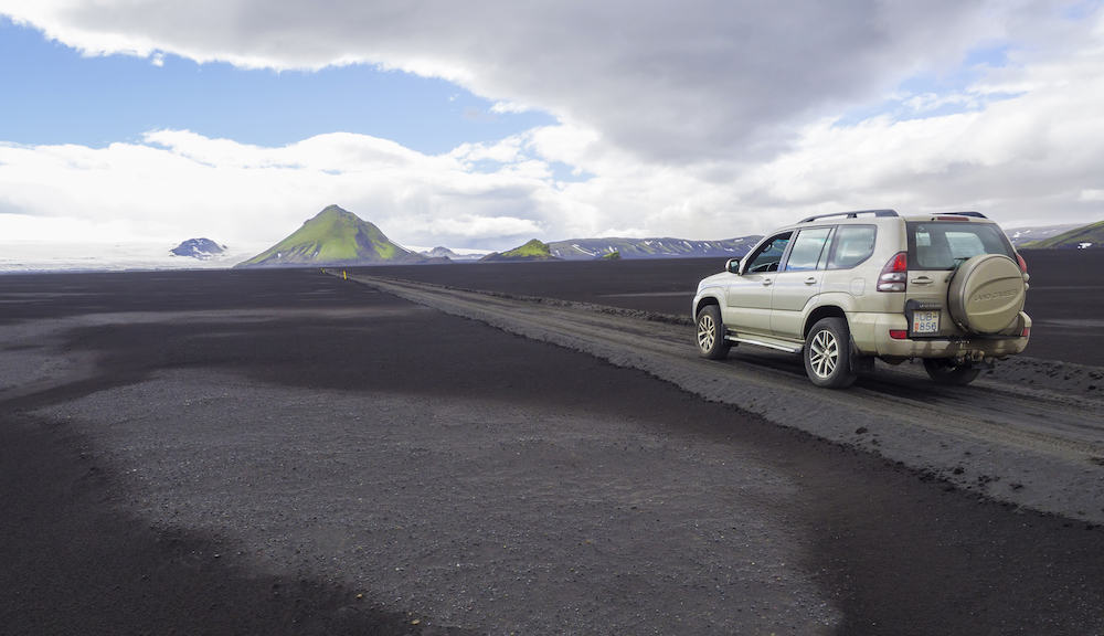 South Iceland, Nature reserve Fjallabak: Off road car Toyota Landcruiser driving on dirt mountain road F210 through black lava sand desert with green Maelifell mountain and myrdalsjokull glacier, blue sky white clouds